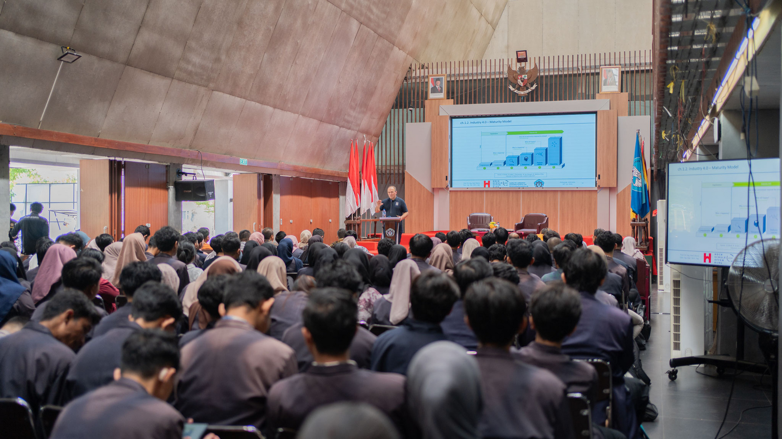 Ein Saal mit Zuhörern und einer Person an einem Rednerpult vor einer Leinwand